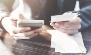 Hands hold a smartphone and receipts, with more receipts on a wooden table