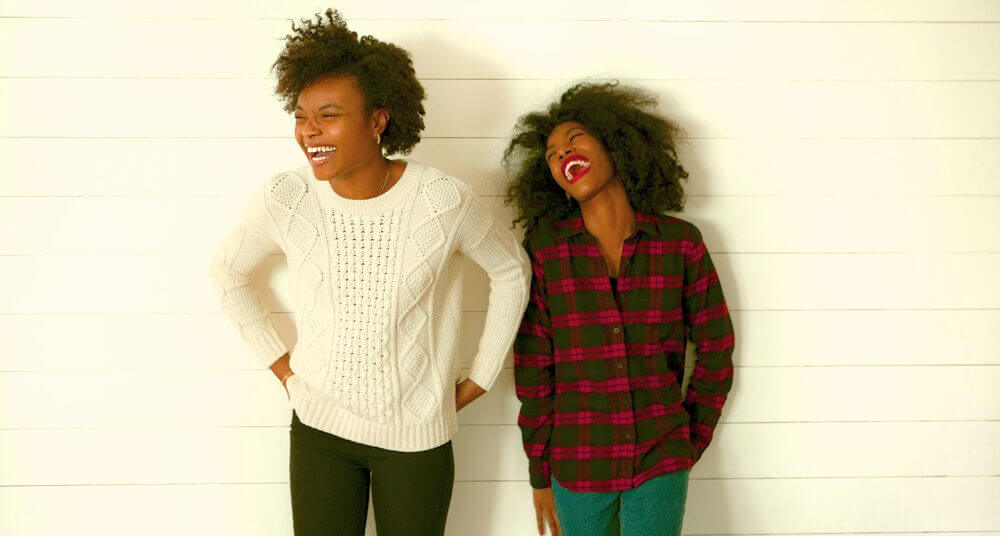 Two young women laughing, one in a white sweater, the other in a red and black plaid shirt