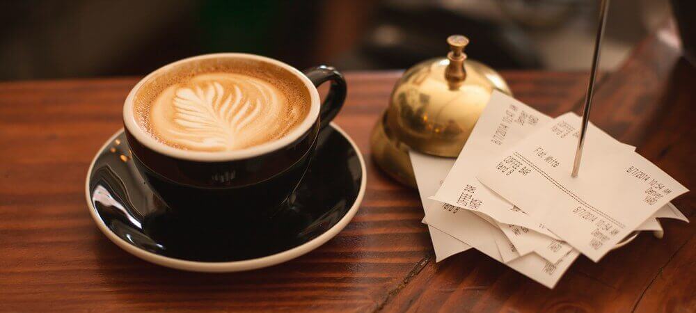 A latte, a brass bell, and a stack of receipts on a wooden counter