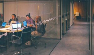 Three people in a modern office meeting room with laptops, seen through a glass wall