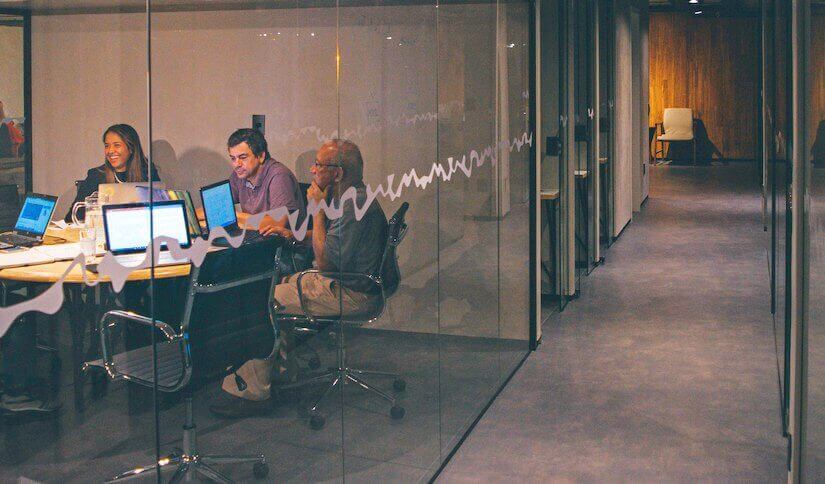 Three people in a modern office meeting room with laptops, seen through a glass wall