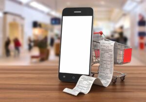 Smartphone displaying a blank screen, a miniature shopping cart, and paper receipts on a wooden surface