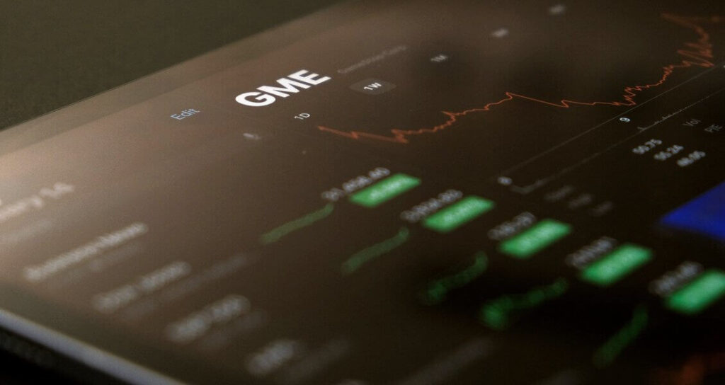 A tablet displays a stock chart for GME, showing a red line graph and green bar charts