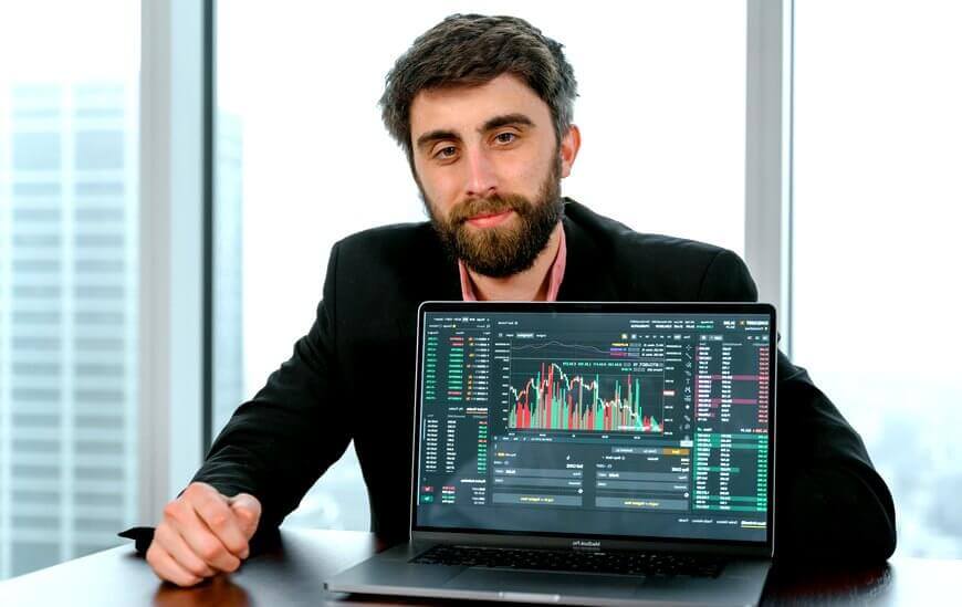 Man with beard and suit smiles at camera, laptop with financial charts on desk