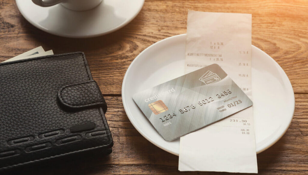 A credit card and receipt on a plate next to a wallet and coffee cup on a wooden table