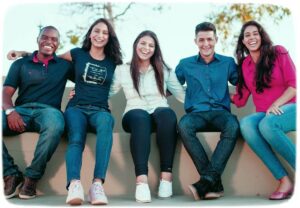 Five diverse young adults smiling, sitting on a bench, arms linked, enjoying each other's company