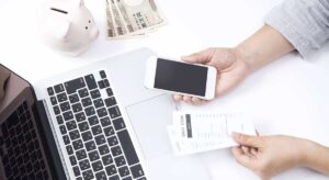 Hands hold a smartphone and a "DRUG STORE" receipt next to a laptop, piggy bank, and yen notes