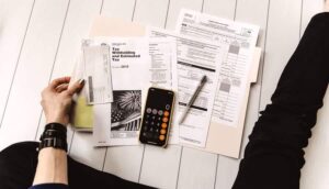 Person preparing taxes with forms, phone calculator, and pen on a white wooden floor