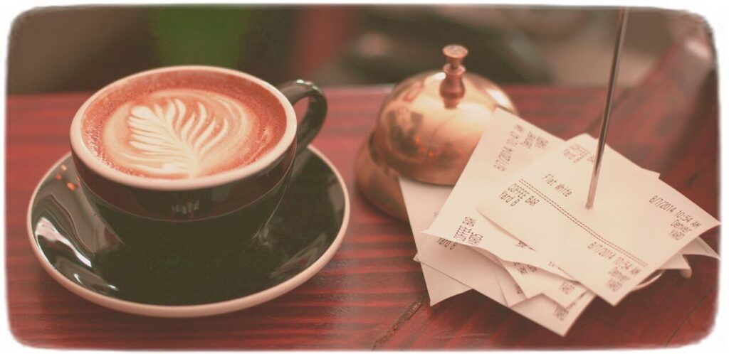 Latte art in a cup next to a stack of receipts on a spike, with a bell behind
