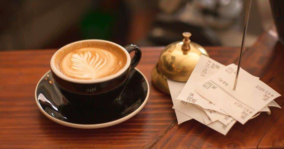 Latte, brass bell, and stack of paper receipts on a wooden counter