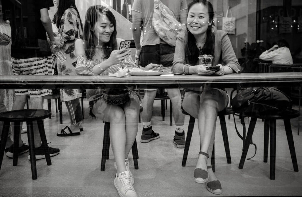 Two women smile and look at phones in a cafe, reflecting a busy street scene