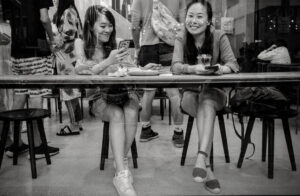 Two women smile and look at phones in a cafe, reflecting a busy street scene