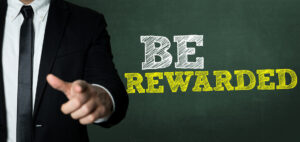 Man in a suit pointing at "BE REWARDED" written in chalk on a green chalkboard