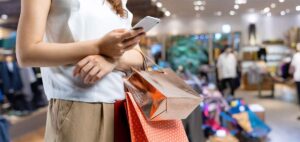 Woman in a store holding shopping bags and checking her phone, likely comparing prices or deals
